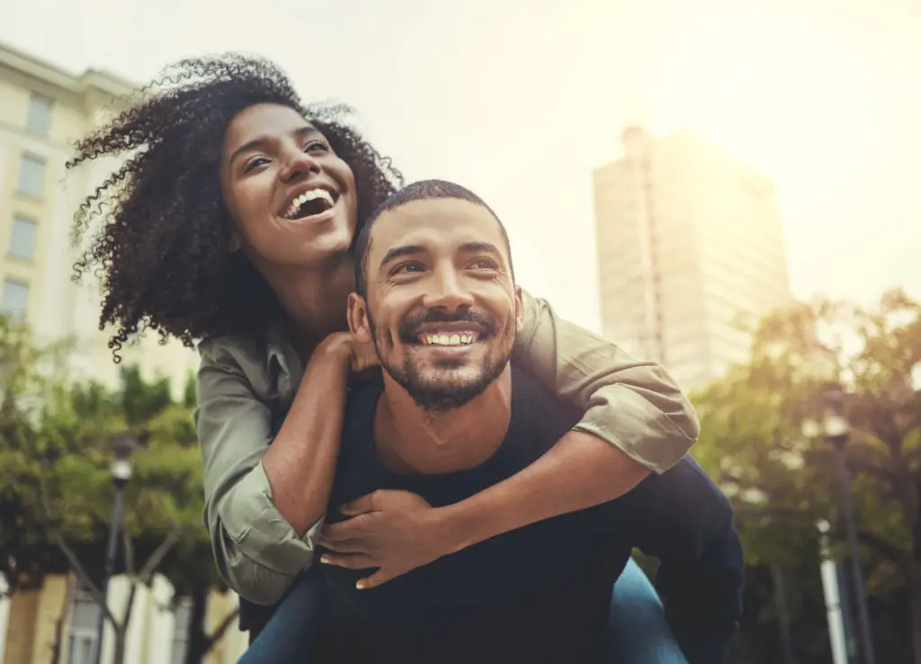 A laughing couple outdoors in the sun, she hugging him from behind.