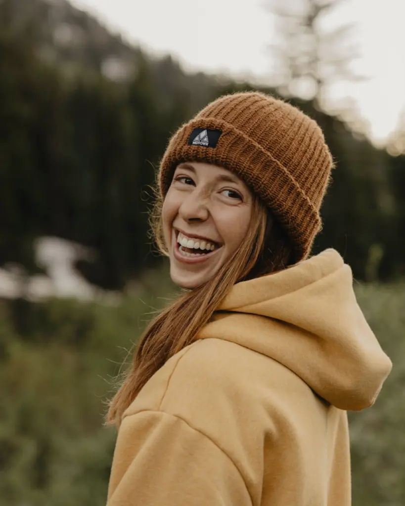 Jo Coleman, laughing over her shoulder on a forest trail, in a knit beanie and a mustard hoodie.