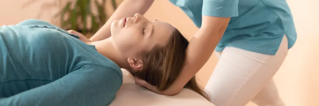 A practitioner gently cradles a relaxed woman’s head and neck as she lies on a treatment table, eyes closed.